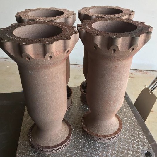 Four additively manufactured combustion chambers with scalloped flanges standing on a build plate.
