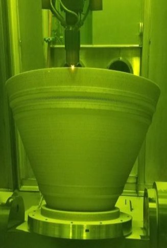 A large nozzle mid-build beneath a deposition head, seen through green laser-safety glass.