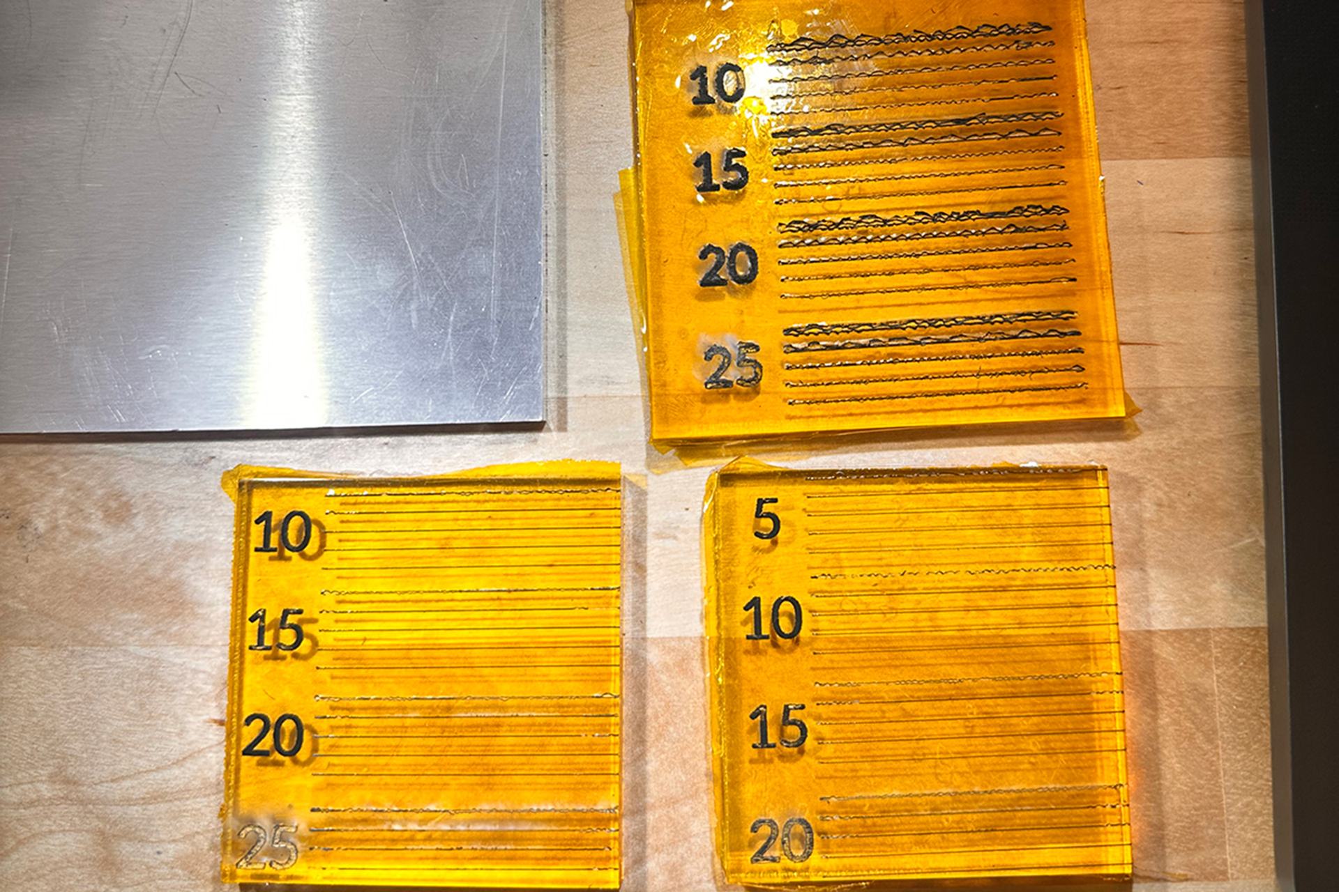 Three Kapton test pieces with groups of dark laser lines numbered 5 to 25, beside a blank piece and an aluminum plate